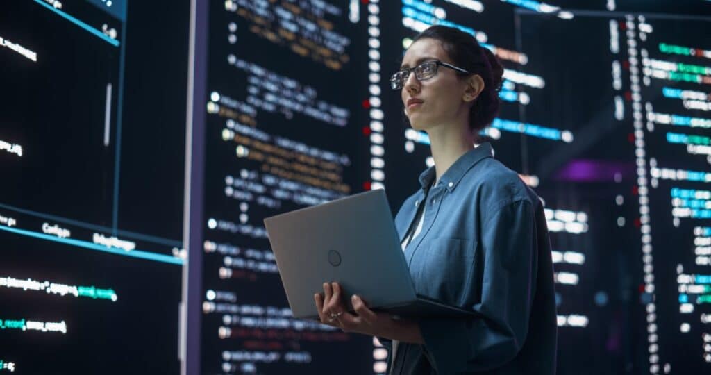 A programmer with a laptop stands in front of walls of code